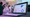 A woman is seen in a hospital bed behind a computer displaying a spreadsheet.